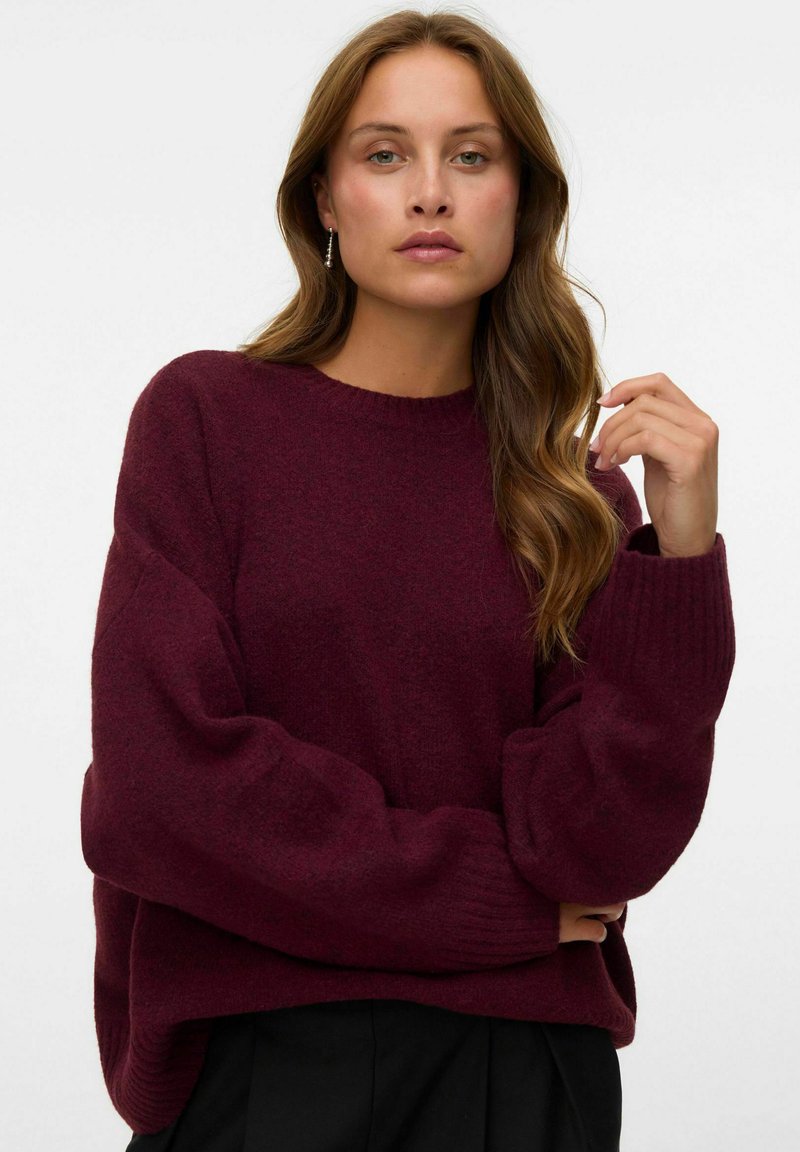 Femme aux longs cheveux ondulés portant un pull ample bordeaux foncé et un pantalon noir, debout devant un fond clair uni.