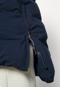 Granatowa kurtka puchowa o teksturowanym wykończeniu, z bocznym zamkiem błyskawicznym i elastycznymi mankietami dla lepszego dopasowania. Widoczne lekkie ocieplenie.