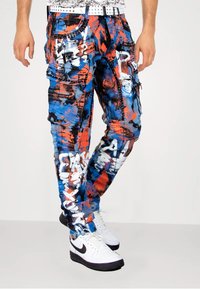 Cargo-Hosen mit einem Blau-, Orange- und Weißfarbspritzdesign, mehreren Reißverschlusstaschen und Nietenakzenten.