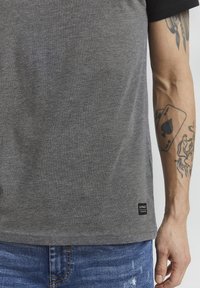Camiseta gris con mangas negras, hecha de tela suave, con una pequeña etiqueta de la marca en el dobladillo, combinada con jeans azules desgastados.