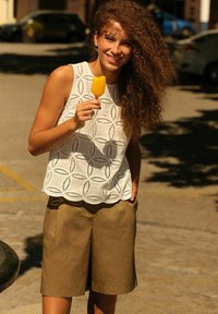 Moteris su garbanotais plaukais šypsosi, laiko geltoną ledinuką, vilki baltą raštuotą bealkūnį marškinėlį ir rudus šortus lauke sausą, saulėtą dieną.