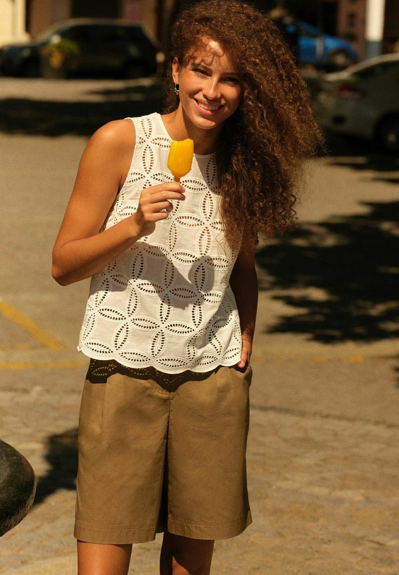 Moteris su garbanotais plaukais šypsosi, laiko geltoną ledinuką, vilki baltą raštuotą bealkūnį marškinėlį ir rudus šortus lauke sausą, saulėtą dieną.