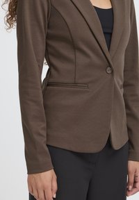 Brown tailored blazer with a single button closure, fitted design, smooth texture, and two side pockets, paired with black pants.