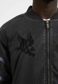 Black bomber jacket featuring an embroidered eagle design on the chest, a zippered front, and ribbed collar. Smooth fabric texture.