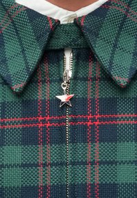 Πουκάμισο καρό με πράσινο και ναυτικό μοτίβο με κόκκινες λεπτομέρειες, ασημένιο φερμούαρ και λαβή σε σχήμα αστεριού. Υφασμάτινη υφή.