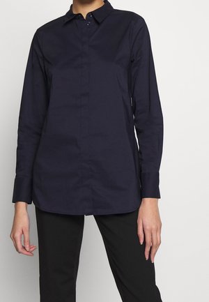 Femme portant une chemise bleu marine à manches longues boutonnée avec un pantalon noir, debout devant un fond clair uni.