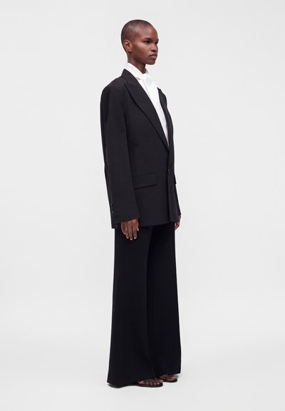 Personne debout vêtue d'un blazer noir oversize, d'un pantalon noir à jambes larges, d'une chemise blanche et de sandales à bout ouvert sur un fond clair uni.