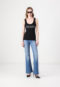 Top sem mangas preto com o logótipo prateado "GUESS", combinado com jeans flare azul claro e sapatos de salto altos pretos, contra um fundo simples.