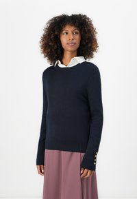 Granatowy sweter dzianinowy z okrągłym dekoltem i prążkowanym dołem. Posiada długie rękawy z złotymi guzikami na nadgarstkach, w połączeniu z białą koszulą z kołnierzykiem.