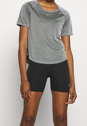 Frau trägt ein graues Nike kurzärmeliges Sportoberteil und schwarze enganliegende Radlershorts, steht mit locker an den Seiten hängenden Armen.