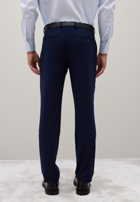 Mann steht mit dem Rücken zur Kamera, trägt navyblaue Anzughose, hellblaues Hemd, schwarzen Gürtel und schwarze Schuhe auf hellem Teppich.