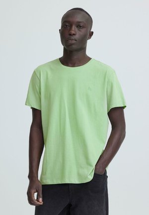 Ung mann med mørk hud som har på seg en lys grønn t-skjorte med korte ermer og svarte bukser, stående mot en ensfarget lys bakgrunn.