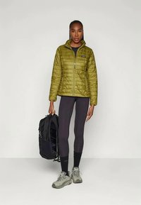 Casaco de cortar vento verde-oliva com fecho éclair à frente e um pequeno logótipo, combinado com leggings pretas e uma mochila preta. Usado com ténis cinzas.