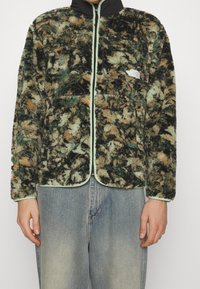 Fleecejacke in Grün, Braun und Schwarz mit weicher Textur, mintgrünen Reißverschluss und Logo auf der linken Brust; verfügt über zwei Fronttaschen.