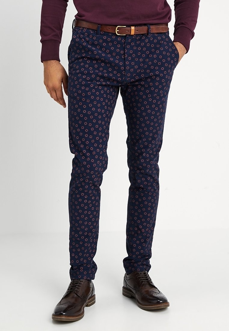 Man in een donkerblauwe slim-fit broek met een klein rood geometrisch patroon, een leren bruine riem en donkerbruine brogueschoenen.