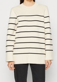 Jersey crema con rayas horizontales negras, cuello redondo y mangas largas. Textura suave y ajuste relajado, combinado con vaqueros oscuros.