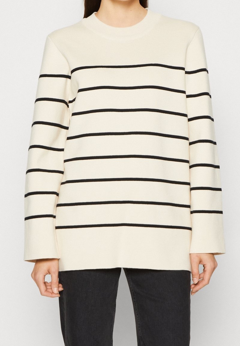 Jersey crema con rayas horizontales negras, cuello redondo y mangas largas. Textura suave y ajuste relajado, combinado con vaqueros oscuros.