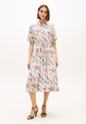 Kvinde iført en midtlang, kortærmet kjole med en taljesamling og abstrakt pink og sort dyreprint, kombineret med olivengrønne højhælede sandaler.