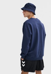 Donkerblauw sweatshirt met een relaxte pasvorm, voorzien van ribgebreide boorden en onderrand. Gecombineerd met zwarte shorts met witte chevronpatronen.
