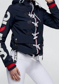 Marinblå vattentät jacka med vita togglestängningar, en röd dragkedja och en stor vit logotyp på armen. Har en design med insnörd midja.