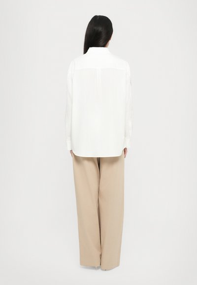Camisa blanca con cuello clásico, manga larga y detalle cortado en la parte posterior, combinada con pantalones anchos beige. Textura de tela suave.