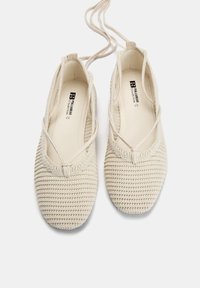 Beige Häkel-Ballerinas mit überkreuzten Riemen, strukturiertem Obermaterial und runder Zehenpartie, ausgestattet mit einem weichen Stofffutter und minimaler Markierung im Inneren.
