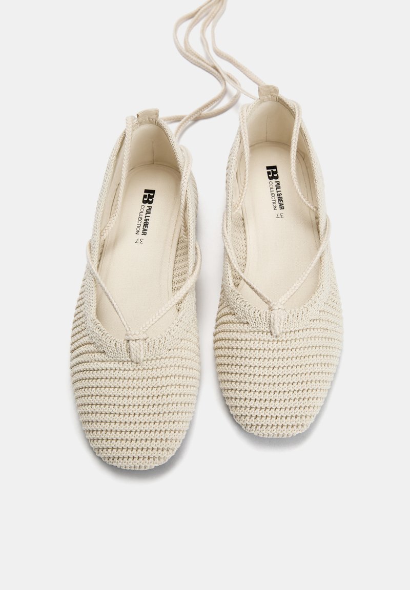 Ballerines en crochet beige avec des brides croisées, surface texturée et bout rond, dotées d'une doublure en tissu doux et d'un logo minimaliste à l'intérieur.