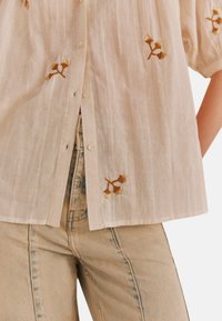 Lys beige bluse med vertikale striber, korte puffærmer og guld broderede blomsteraccenter. Denim jeans med et falmet blåt mønster.