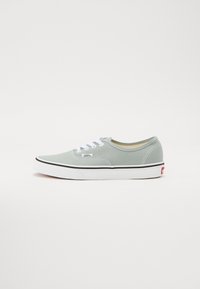 Ljusgrå canvas sneakers med vita snören, svart gummisula och en röd accentlogotyp på hälen. Rund tå och klassisk design.