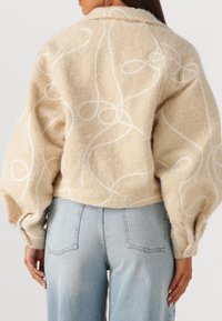 Beige textuurjack met witte draaiende borduursels, met pofmouwen en een kort ontwerp, gecombineerd met lichtblauwe rechte jeans.