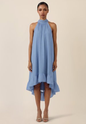 Frau trägt ärmelloses, hellblaues Neckholder-Kleid mit hohem, gerüschtem Saum vorne und beige offenen High-Heels, steht vor schlichtem Hintergrund.