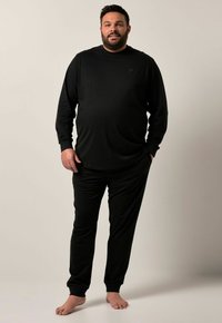 Hombre con barba vistiendo una camisa de manga larga negra y pantalones, de pie descalzo frente a un fondo neutro con una mano en el bolsillo.
