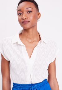 Blouse blanche à manches courtes avec une texture ondulée, col à boutons et décolleté en V. Assortie à un pantalon bleu à taille haute avec cordon de serrage.