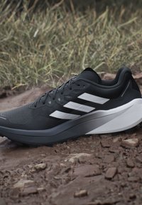 Scarpa da corsa bianca e nera con tre strisce bianche, appoggiata su un terreno roccioso con erba sullo sfondo.