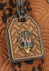 Etichetta in pelle intrecciata marrone con un design di pesce colorato con finitura metallica, accentuata da gemme multicolori. Sfondo testurizzato.