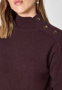 Pull bordeaux avec un col montant à côtes, doté de trois boutons décoratifs marron sur une épaule et d'une finition douce et texturée.