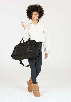 Zwarte weekendtas met twee draagbanden, met een gestructureerd oppervlak en een zijvak. Model in een lichte top en leggings, met tan laarzen.