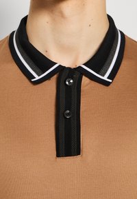 Gros plan sur un polo marron avec un col noir comportant des rayures blanches et deux boutons noirs sur la patte.