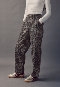 Cargo-Hosen mit Leopardenmuster in gedämpften Tönen, ausgestattet mit zwei Fronttaschen und einem lockeren Schnitt. Kombiniert mit flachen Sneakern in Beige mit bordeauxfarbenen Akzenten.