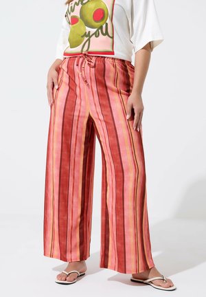 Personne portant un pantalon à jambes larges avec des rayures verticales dans des tons rouges, roses et orange, associé à une chemise blanche et des tongs blanches.