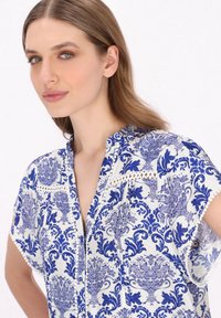 Hvid bluse med et blåt blomsterdesign, med mandarin krave, korte ærmer og blonde detaljer langs halsudskæringen og skuldersømmerne.
