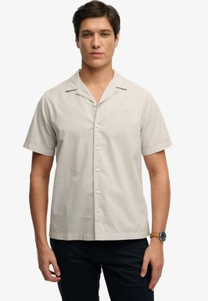 Ung mand iført en beige kortærmet skjorte med knapper og mørke bukser, stående med et neutralt udtryk mod en hvid baggrund.