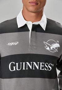 Dark gray rugby shirt with horizontal black and light gray stripes. Features a white collar and printed logos for Guinness and a toucan.