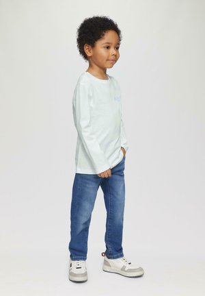 Lichtblauwe langemouwen T-shirt met een kleine grafische print, gecombineerd met donkerblauwe spijkerbroeken en witte sneakers met grijze accenten.