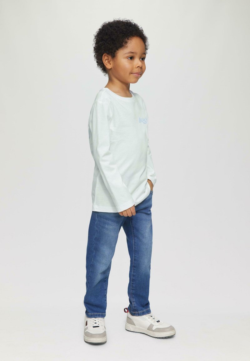 Lichtblauwe langemouwen T-shirt met een kleine grafische print, gecombineerd met donkerblauwe spijkerbroeken en witte sneakers met grijze accenten.