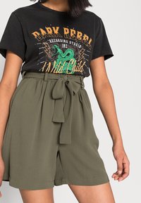 T-shirt nera con grafica di un serpente verde e testo arancione, abbinata a pantaloni corti a vita alta color verde oliva con cintura annodata.