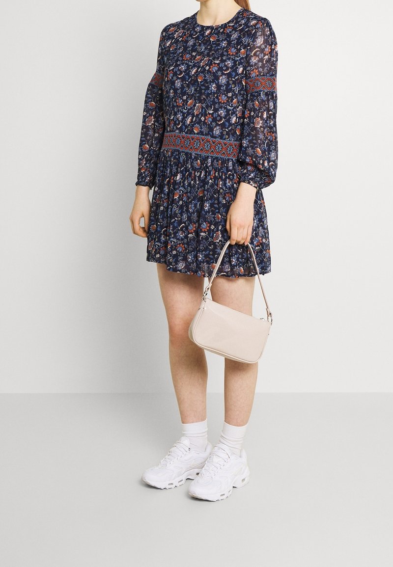 Femme portant une robe florale bleu marine avec des accents rouges, des baskets blanches et des chaussettes, tenant un petit sac à main beige à ses côtés.
