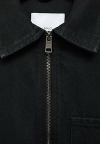 Črna jakna z zadrgo na sprednjem delu, s koničastim ovratnikom in eni prsni žep. Narejena iz trdnega blaga z vidnimi šivi.