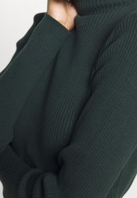 Tummanvihreä ribbikuvioinen neulepaita, jossa on korkea kaulus ja pitkät hihat, joissa on teksturoitu pinta. Kangas näyttää pehmeältä ja joustavalta.