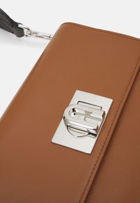 Brązowa skórzana kopertówka z polerowanym srebrnym zamknięciem, o prostokątnym kształcie i gładkich krawędziach. Dołączony regulowany czarny pasek.